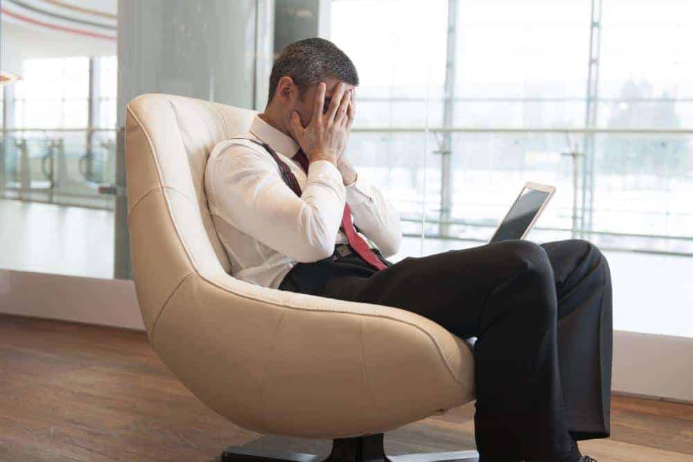 A businessman sits with a laptop on his legs and with both hands covering his face, looking stressed out.
